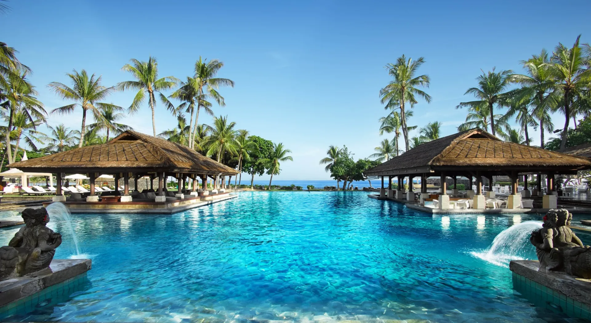 Turquoise resort pool framed by palms and thatched pavilions, opening to the sea